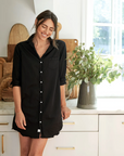 A woman in a black Mary Classic Shirtdress from Frank & Eileen smiles warmly, standing in the bright kitchen of her Scottsdale, Arizona bungalow with wooden accents and a vase of greenery on the countertop beside her.