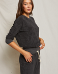 A woman in a Perfectwhitetee Saylor Loop Terry Sweatshirt and matching sweatpants stands against a light beige background in a bungalow, her hand on her hip, looking at the camera with a subtle smile.