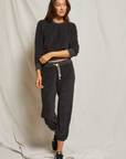 A woman stands against the beige backdrop of a bungalow, wearing a casual dark gray Saylor Loop Terry Sweatshirt and matching joggers with white drawstrings from Perfectwhitetee. She smiles lightly, her left hand resting on her hip.