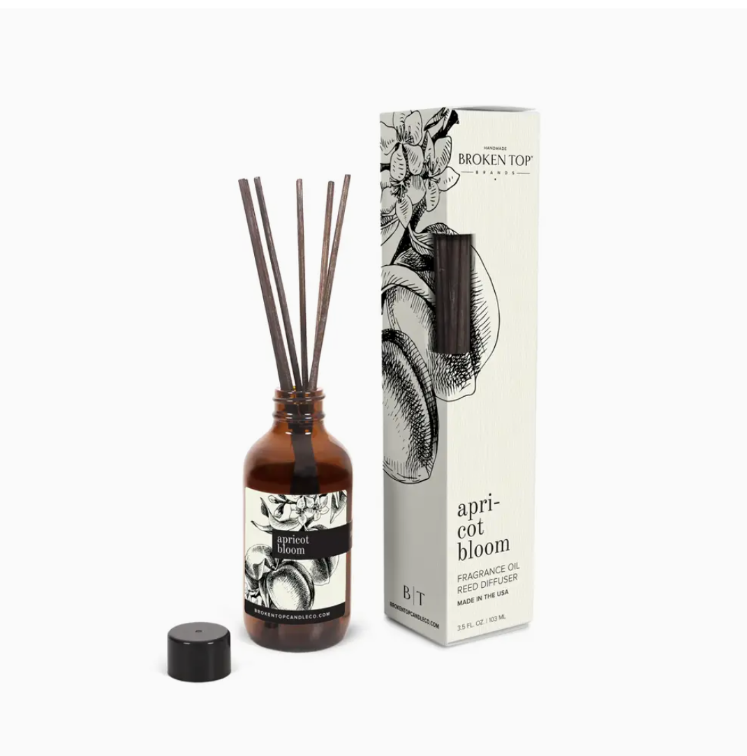 A Faire reed diffuser in an amber glass bottle labeled &quot;apricot bloom&quot; with several dark sticks inserted, positioned next to its floral-decorated packaging box, perfect for home decor.