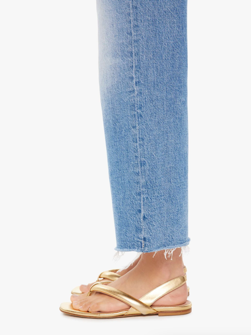 Side view of a person&#39;s lower leg wearing Mother&#39;s Maven Ankle Fray jeans and gold strappy sandals, standing against a white background.