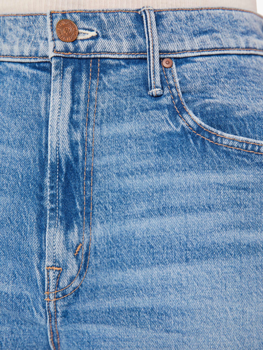 Close-up view of Mother&#39;s Maven Ankle Fray denim skirt featuring detailed stitching, front pockets, and bronze-colored button and rivet. The texture of the denim is clearly visible.