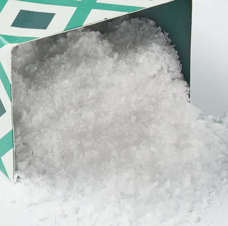 A mound of Flaky Maldon Sea Salt spilling out from a partially opened Faire green and white cardboard box on a white background.