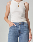 A woman in a sleeveless white ribbed racerback Isabel Rib Tank by Citizens Of Humanity/AGOLDE and blue jeans, with one hand casually tucked into the pocket. She wears a simple golden necklace. The background is plain and light-colored.