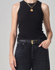 A person wearing a black sleeveless Isabel Rib Tank by Citizens Of Humanity/AGOLDE and dark jeans with a black belt and a gold buckle, accessorized with a gold pendant necklace. The setting is against a neutral background.
