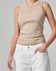 A person wearing a Citizens Of Humanity/AGOLDE beige sleeveless ribbed racerback tank and white jeans, accessorized with a gold pendant necklace, standing against a grey background. Only the torso and hands are visible.