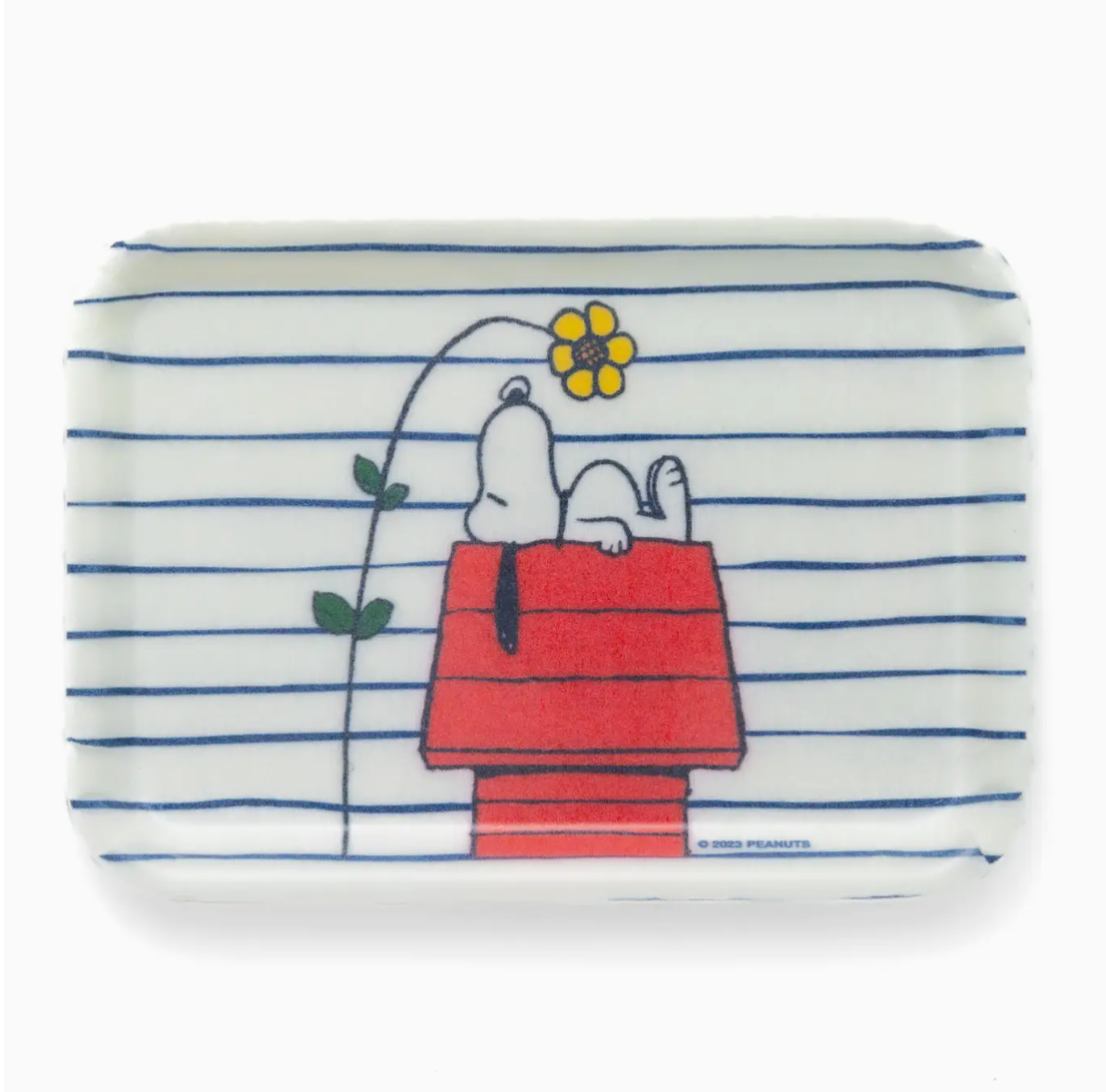A rectangular soap bar featuring an illustration of Snoopy sitting on a red bungalow with a yellow flower in his mouth, set against a background with blue horizontal stripes - Faire's Peanuts Tray.