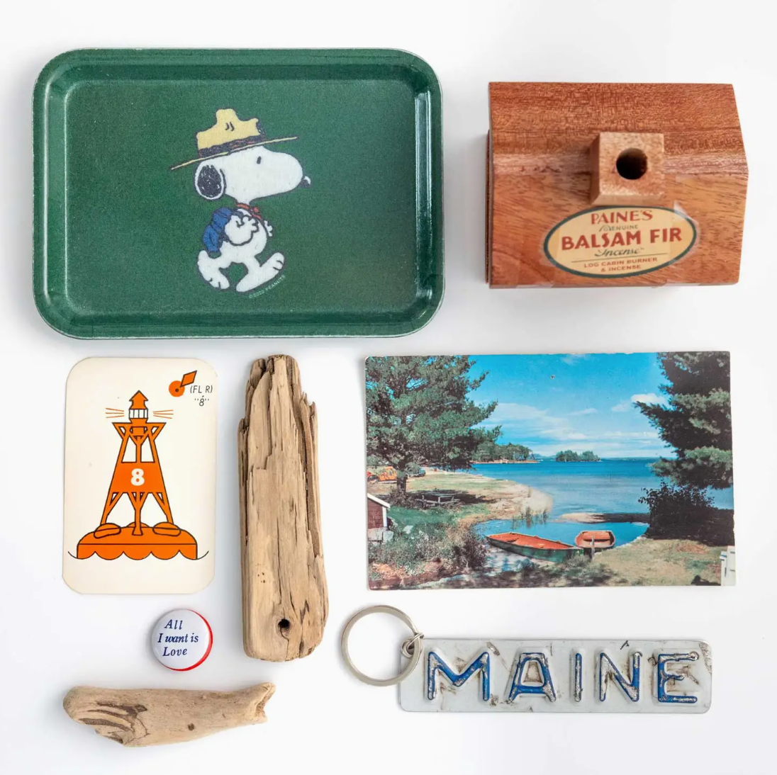 Various mementos arranged on a white background: a tray with a Peanuts design from Faire, a wooden box labeled "Paines Balsam Fir," a playing card featuring a lighthouse, a driftwood piece from Scottsdale Arizona, a postcard of a seaside view, and a keychain spelling.