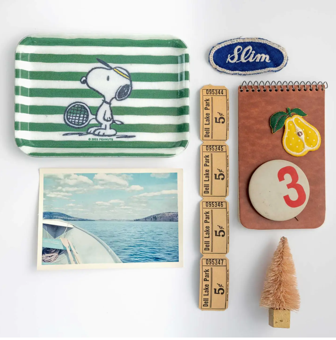 Assorted items on a white background: a Peanuts Tray with Snoopy playing tennis, a "Slim" patch, a photo of a boat at sea near Scottsdale, Arizona, a folded ruler, a notepad with a lemon design in front of a bungalow, soap with number 3.