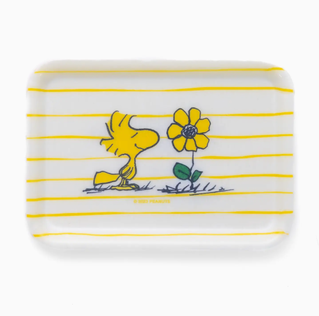 A rectangular soap bar featuring a design with the Peanuts character Woodstock next to a yellow flower, set against a white background with yellow horizontal stripes in a bungalow style, from the Faire Peanuts Tray.