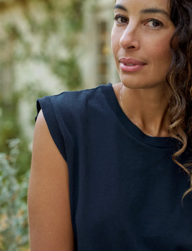 Close-up of a woman with curly hair, wearing a Frank &amp; Eileen AIDEN Vintage Muscle Tee HERITAGE JERSEY in dark blue, slightly smiling at the camera in a leafy outdoor setting near a Scottsdale Arizona bungalow.
