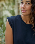 Close-up of a woman with curly hair, wearing a Frank & Eileen AIDEN Vintage Muscle Tee HERITAGE JERSEY in dark blue, slightly smiling at the camera in a leafy outdoor setting near a Scottsdale Arizona bungalow.