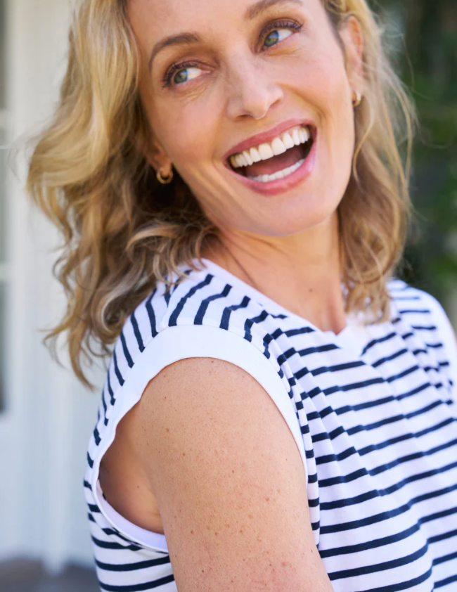 A joyful middle-aged woman with blonde hair, laughing and looking at the camera, wearing a Frank &amp; Eileen AIDEN Vintage Muscle Tee HERITAGE JERSEY outdoors in Scottsdale, Arizona with a blurred green background.