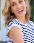 A joyful middle-aged woman with blonde hair, laughing and looking at the camera, wearing a Frank & Eileen AIDEN Vintage Muscle Tee HERITAGE JERSEY outdoors in Scottsdale, Arizona with a blurred green background.