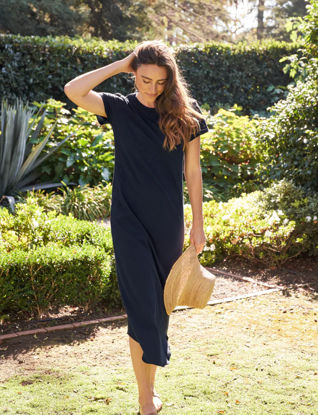 A woman walks through a sunny garden in Scottsdale, Arizona, holding a straw hat, dressed in a dark blue jumpsuit, with her eyes cast downwards and a contemplative expression wearing the Frank & Eileen Harper Perfect Tee Maxi.