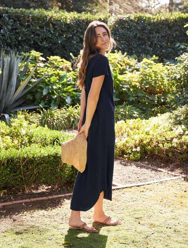 A woman in a navy blue Frank & Eileen Harper Perfect Tee Maxi dress and sandals holding a straw hat walks on a garden path by a bungalow with lush greenery and sunlight filtering through in Scottsdale, Arizona.