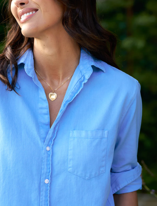 Close-up of a smiling woman wearing a Frank &amp; Eileen Mary Classic Shirtdress and a heart-shaped pendant necklace, standing outdoors with greenery in the background in Scottsdale, Arizona.