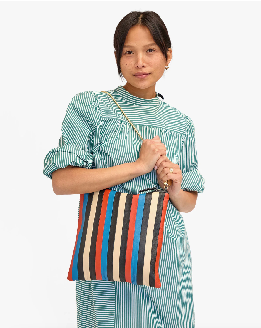 A woman wearing a blue and white striped dress holds a Clare Vivier Foldover Clutch w/ Tabs Multi Stripes Nappa, featuring red, blue, black, and cream vertical stripes, posing in Arizona-style against a white background.