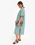A woman in a striped dress and black sandals smiles slightly while holding a colorful Clare Vivier Foldover Clutch w/ Tabs Multi Stripes Nappa, standing against a white background, exuding effortless Arizona style.