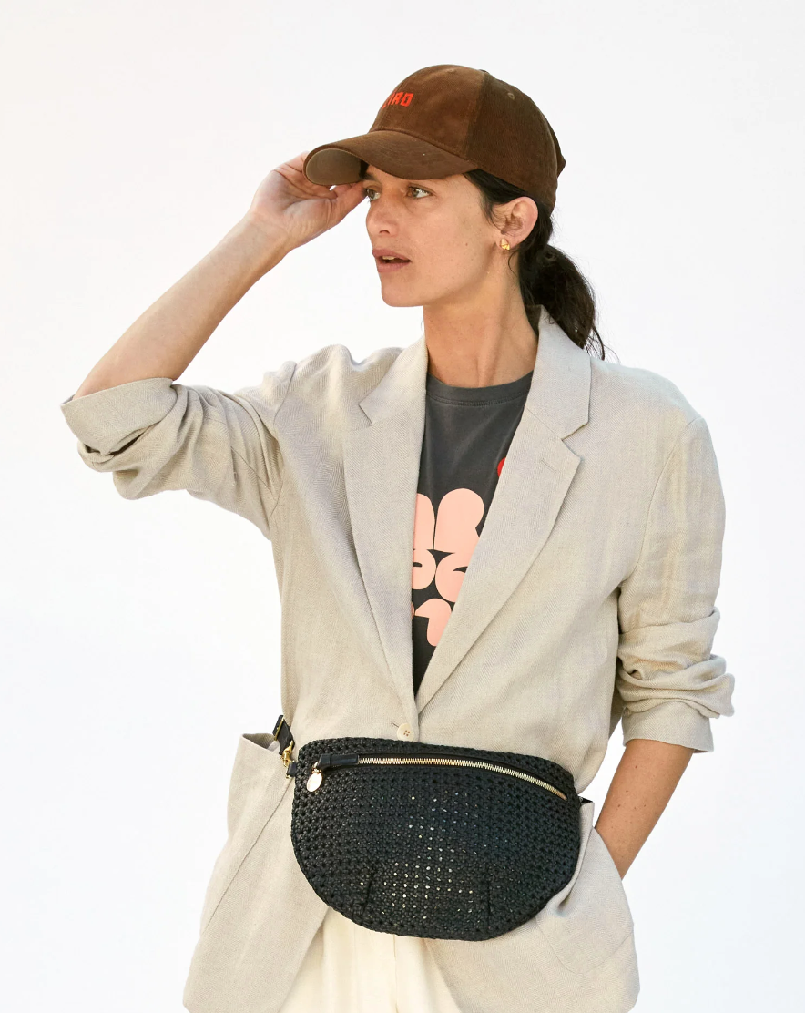 A woman in a casual chic outfit with a beige blazer and white pants, wearing a brown cap and carrying a Clare Vivier Grande Fanny crossbody bag, looking away thoughtfully in Arizona.