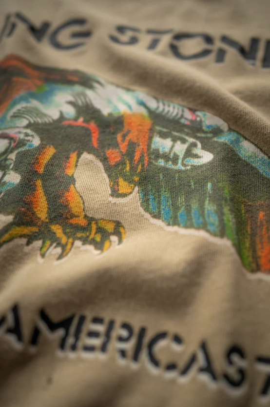 Close-up of a beige classic tee with a colorful graphic print featuring a tiger and text. The top section reads &quot;THE STONE&quot; and the bottom &quot;AMERICAS.&quot; The Rolling Stones Americas &#39;75 Tour tee by Made Worn is slightly out of focus near the edges.