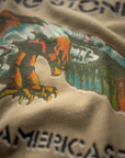 Close-up of a beige classic tee with a colorful graphic print featuring a tiger and text. The top section reads "THE STONE" and the bottom "AMERICAS." The Rolling Stones Americas '75 Tour tee by Made Worn is slightly out of focus near the edges.