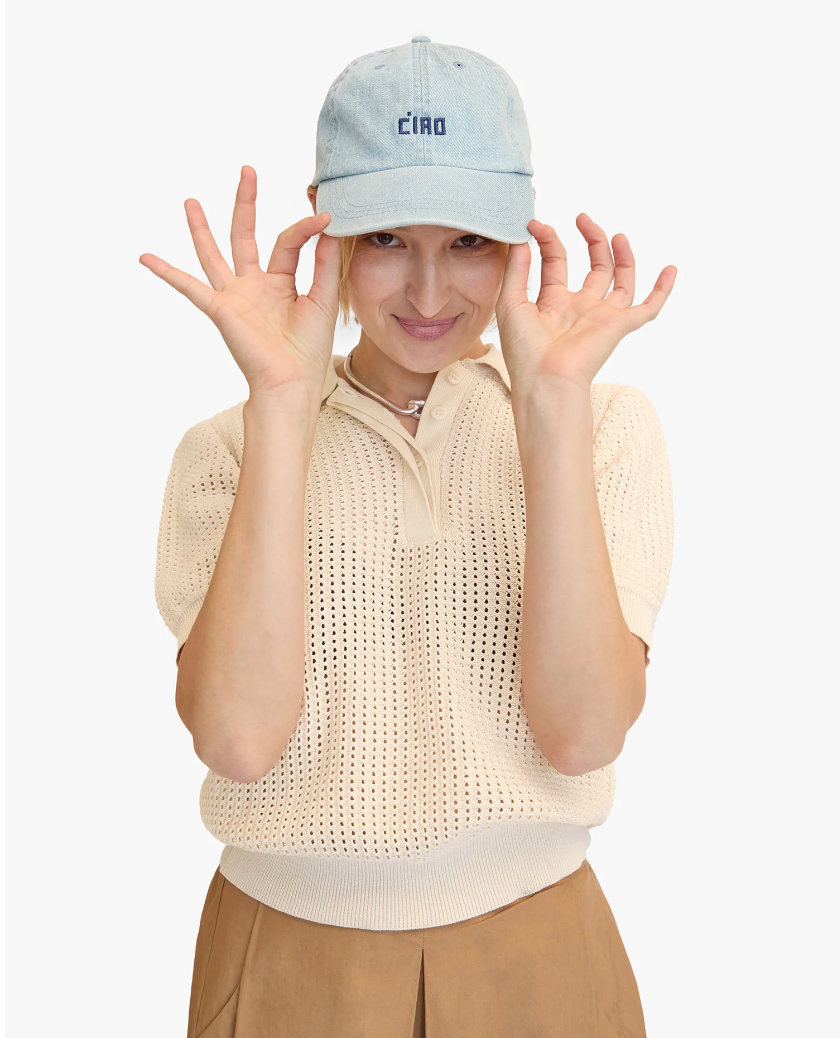 A smiling woman in a Baseball Hat Petit Block Ciao Denim cap and beige sweater making an "OK" gesture with both hands near her eyes. She wears a white collared shirt underneath and stands against a white background, embodying the casual Arizona style from Clare Vivier.