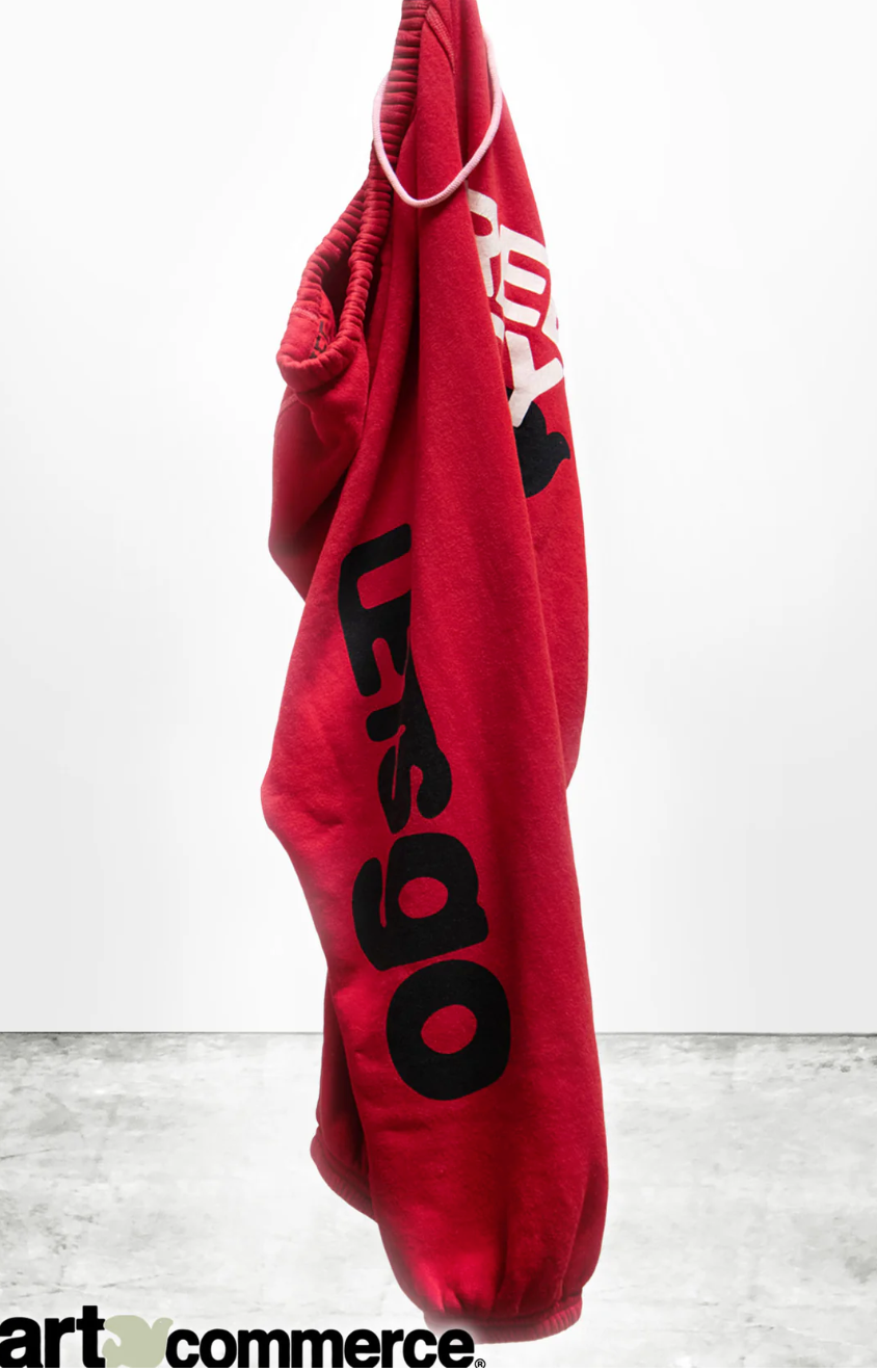 A CIRCA'99 OG LETSGO OLDSCHOOL POLYBLEND/FLUFF sweat pant by Free City with hand-screen printed black text hanging against a white wall, partially obscured by being folded over a hanger.