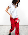 A woman wearing red CIRCA'99 OG LETSGO OLDSCHOOL POLYBLEND/FLUFF sweatpants with the word "FRESH" and a plain white Free City (sparrow, LLC) T-shirt is standing against a plain white background, her hair in mid-movement.