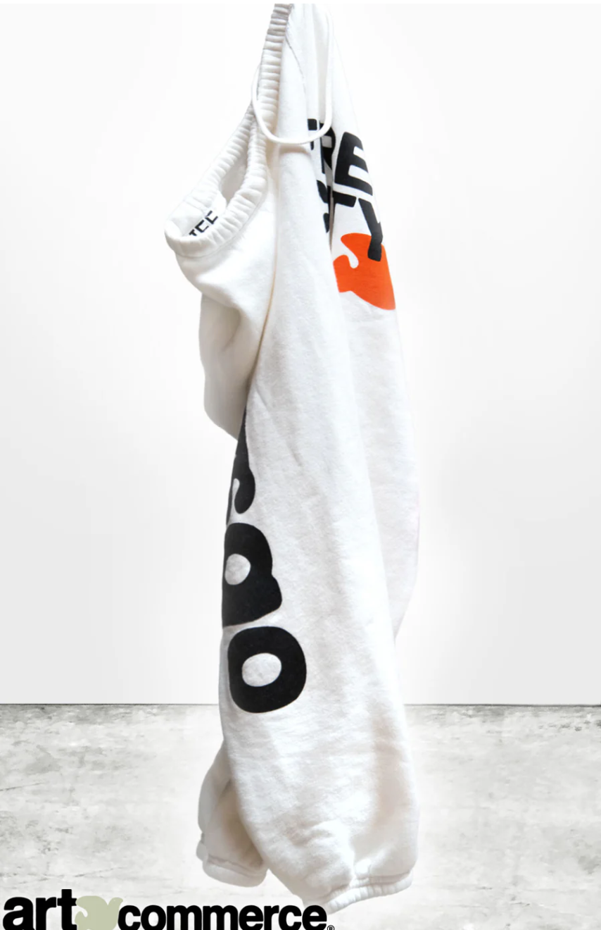 A white CIRCA'99 OG LETSGO OLDSCHOOL POLYBLEND/FLUFF sweat pant with black and orange lettering, hand-screen printed, hung on a hook against a plain, light grey background. The text on the hoodie is partially visible and stylized.