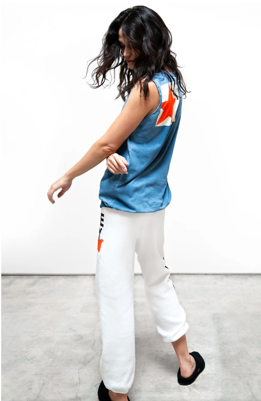 A woman with dark, curly hair is wearing a blue sleeveless top with a red star and white Free City (sparrow, LLC) CIRCA&#39;99 OG LETSGO OLDSCHOOL POLYBLEND/FLUFF sweat sweatpants, walking to her left in a bright, neutrally colored space.