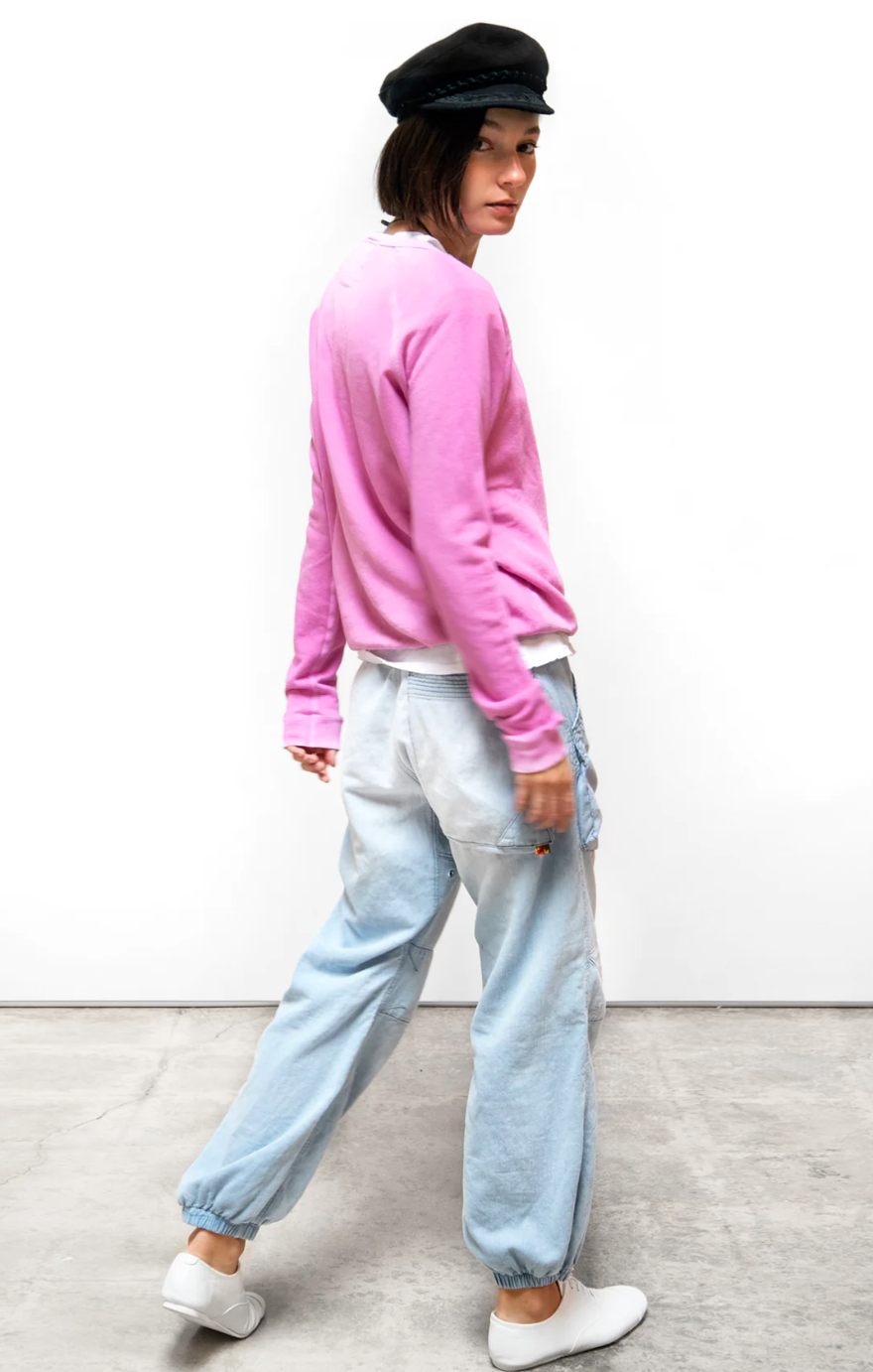 A woman models casual fashion, looking over her shoulder. She wears a pink sweatshirt, FLAP/SNAP INDIGO pants from Free City (sparrow, LLC), white sneakers, and a black beret.