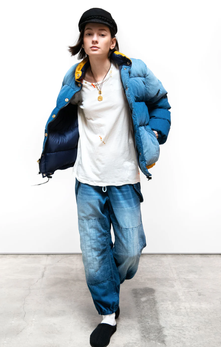A young person in stylish, layered streetwear: an oversized blue puffer jacket over a white t-shirt, FLAP/SNAP INDIGO pants from Free City (sparrow, LLC), a black beanie, and beige bucket hat, posing against a white background.