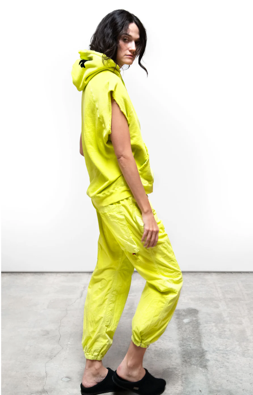 A woman stands sideways wearing a bright yellow unisex CUTOFF SUPERYUMM BIGGIE hoodie from Free City (sparrow, LLC) with a playful expression, against a plain white background. She has dark hair and simple black footwear.