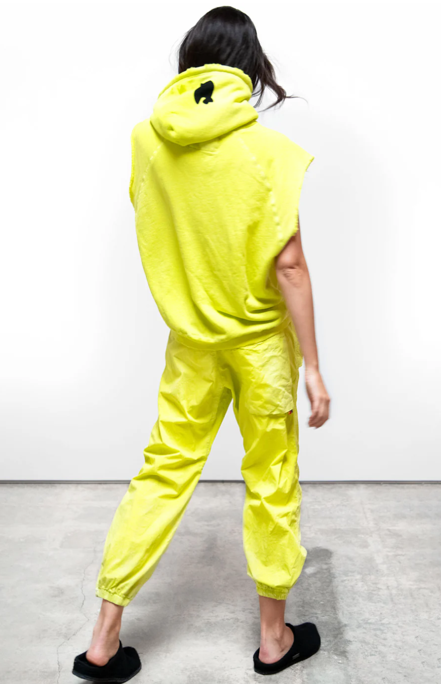 A woman in a striking neon yellow outfit, viewed from behind. She wears a unisex Free City (sparrow, LLC) CUTOFF SUPERYUMM BIGGIE hoodie and matching pants, with her hood up, revealing a small black logo on the hood.