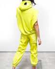 A woman in a striking neon yellow outfit, viewed from behind. She wears a unisex Free City (sparrow, LLC) CUTOFF SUPERYUMM BIGGIE hoodie and matching pants, with her hood up, revealing a small black logo on the hood.