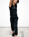 A woman in a casual black outfit with Free City (sparrow, LLC) custom 3d back pocket stretches her right arm over her head, looking down thoughtfully, against a plain white backdrop. She wears slippers and a pendant necklace.