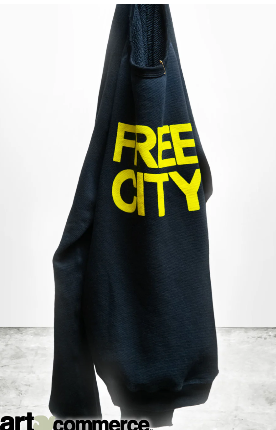 A navy blue FREECITY SUPERYUMM BIGGY RAGLAN hoodie hanging on a hook with the words "FREE CITY" in bold yellow letters across the front, against a gray and white backdrop. This French terry garment is made in USA.
