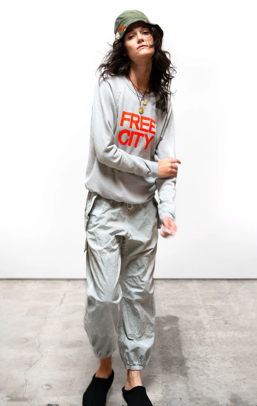 A woman in casual attire, wearing an orange Free City (sparrow, LLC) "FREECITY SUPERYUMM BIGGY RAGLAN" sweatshirt and grey pants, dances with her hair tossed mid-movement against a plain white background.