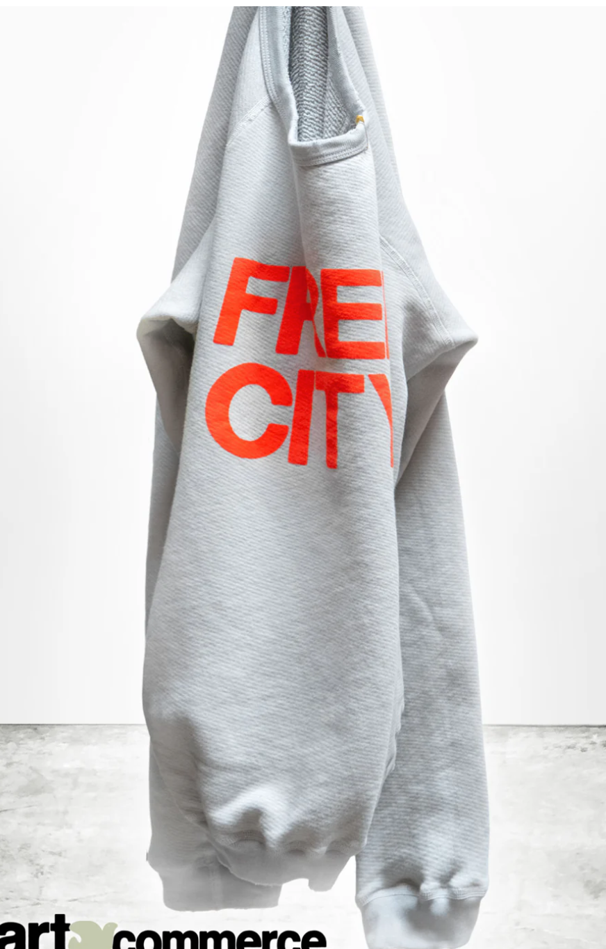 A gray hoodie with bold red text &quot;FREECITY SUPERYUMM BIGGY RAGLAN&quot; hanging against a white wall in Arizona style by Free City (sparrow, LLC).