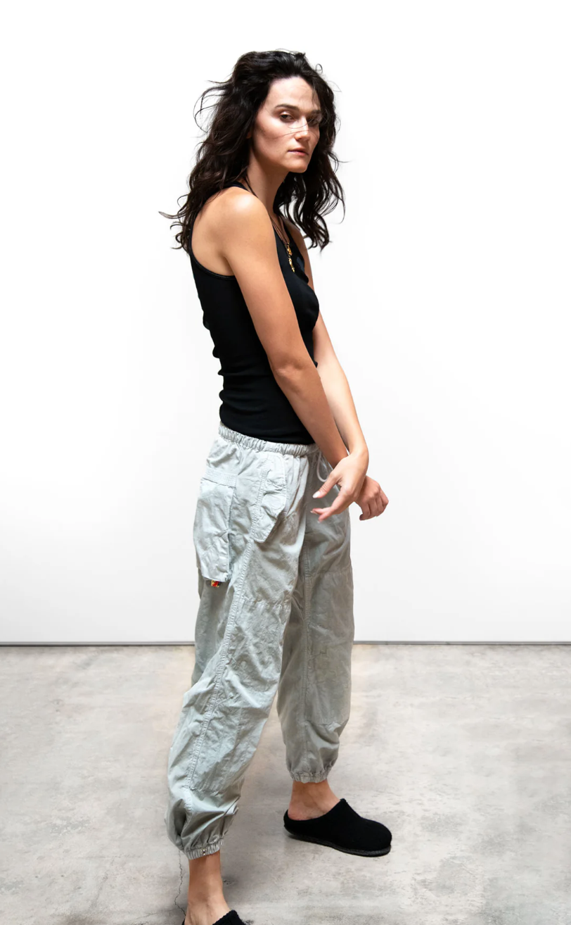 A woman with curly dark hair stands confidently in a Scottsdale studio, wearing a black tank top and light grey OUTOFSIGHT POPLIN/AIR FLAP/SNAP JUMPpant pants by Free City (sparrow, LLC), looking off to the side.