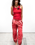 A woman in a red tank top and shiny red OUTOFSIGHT POPLIN/AIR FLAP/SNAP JUMPpant stands confidently against a white background, reminiscent of the minimalist style often found in a Scottsdale, Arizona bungalow. She has dark curly hair and wears black slip-on shoes from Free City (sparrow, LLC).
