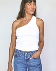 A woman wearing a white Perfectwhitetee Call Me One Shoulder Structured Rib Tank and blue jeans, standing against a plain gray background of a bungalow and looking slightly to her left with a subtle smile.