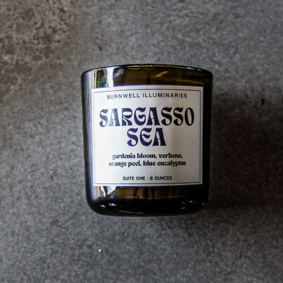 A Barnaby Black Candle jar labeled &quot;Sargasso Sea&quot; with scent notes of gardenia bloom, verbena, orange peel, and blue eucalyptus, displayed on a bungalow-style gray background by Faire.