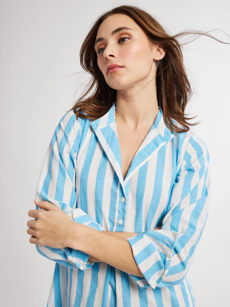 A woman with shoulder-length brown hair wearing a blue and white striped button-up shirt looks thoughtfully to the side on the porch of her Scottsdale, Arizona bungalow, her hair blowing slightly in the breeze wearing a Sadie Long Caftan by Mille.