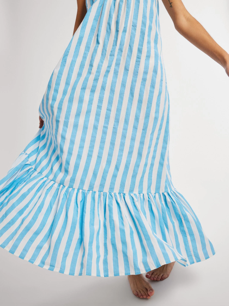A close-up of a Mille Sienna Dress in light blue and white stripes fluttering, with a view of a woman's lower arm and bare feet on a white bungalow background.