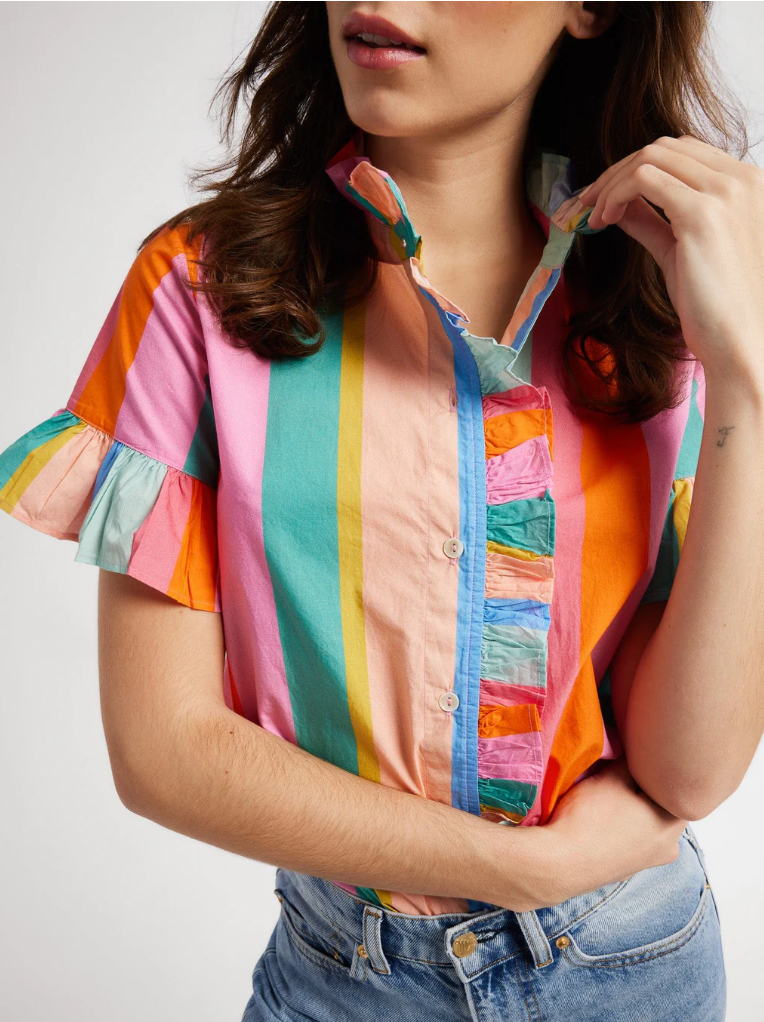 A woman in a vibrant, multicolored striped Mille Vanessa Top with bell-shaped sleeves and blue jeans, partially cropped to show only from her lips downward, with her hand adjusting her collar.