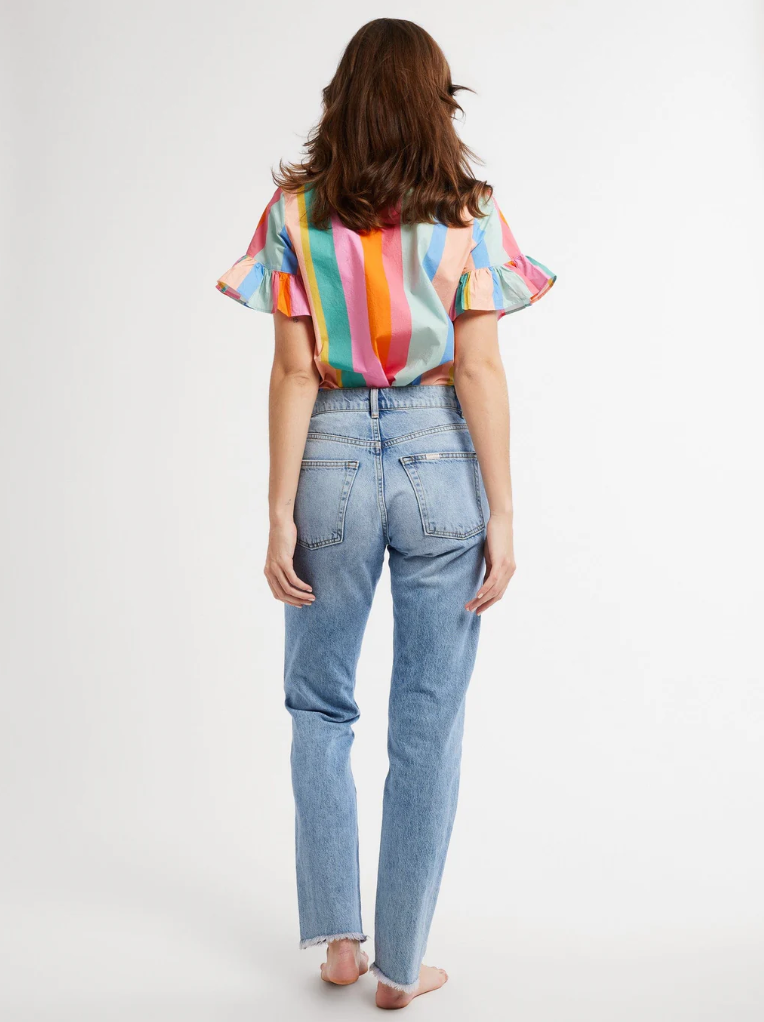 A woman stands facing away from the camera, wearing a colorful striped Vanessa Top from Mille with bell-shaped sleeves and blue jeans, on a plain white background.