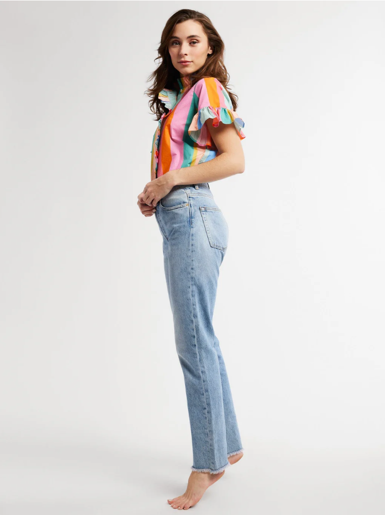 A woman in blue jeans and a Vanessa Top by Mille with cascading ruffles stands with her back slightly turned, looking over her shoulder against a plain background. She is barefoot and her jeans are frayed at the ankles.