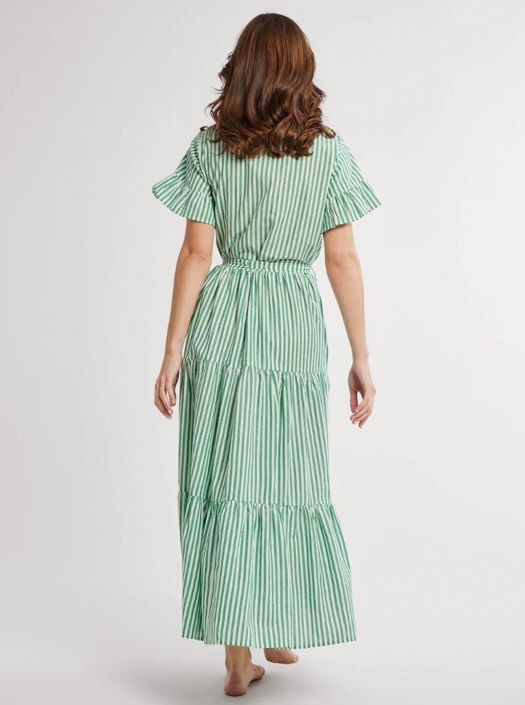 A woman from behind wearing a Mille Victoria Dress, a green and white striped mid-length dress with short sleeves and a cinched waist, standing against the neutral background of a Scottsdale, Arizona bungalow.